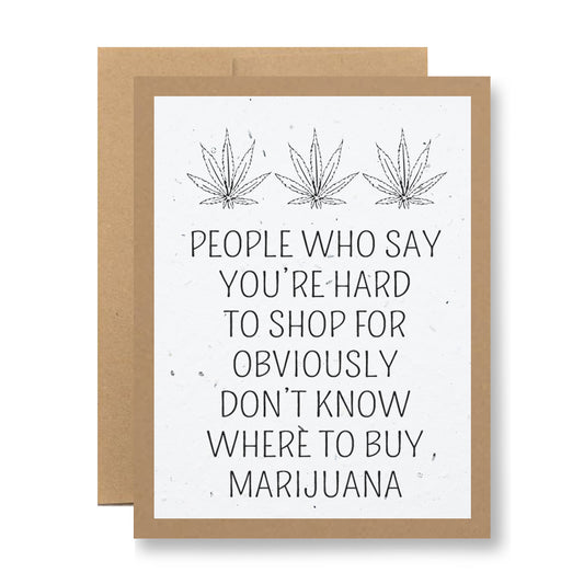 A plantable greeting card from My Store, made with biodegradable seed paper, showcases an illustration of three marijuana leaves at the top. Below, it humorously states: "People who say you're hard to shop for obviously don't know where to buy marijuana." The card is paired with a brown envelope and is designed to blossom into wildflower seeds.