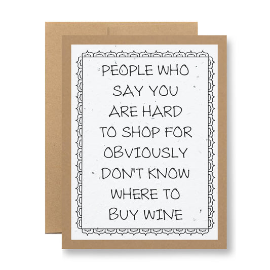 The "Plantable Greeting Card - ...where to buy wine" by My Store is crafted from handmade seed paper and features a humorous message in bold text: "PEOPLE WHO SAY YOU ARE HARD TO SHOP FOR OBVIOUSLY DON'T KNOW WHERE TO BUY WINE." It boasts an intricate border and is set against a plain brown background, ready to blossom into wildflowers.