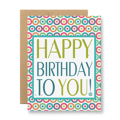 Introducing the "Plantable Greeting Card - Happy Birthday to You!" by My Store. This vibrant birthday card boasts a geometric circle pattern in pink, green, and blue on its background. Made from handmade seed paper, it showcases bold multicolored text proclaiming "Happy Birthday to You!". A partially visible brown envelope accompanies the card.