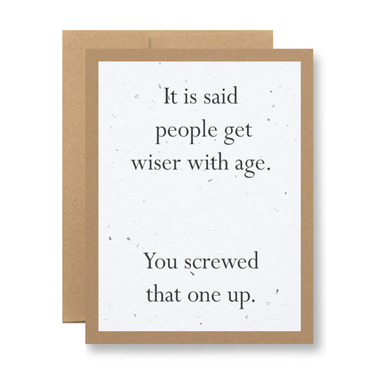 The Plantable Greeting Card from My Store has a brown border and humorously states, "It is said people get wiser with age. You screwed that one up." A plain brown envelope crafted from seed paper is slightly visible behind the card, designed to grow into wildflower seeds.