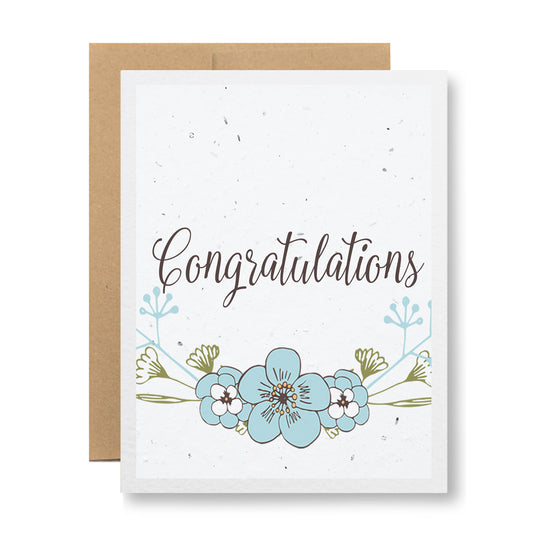 The My Store "Plantable Greeting Card - Congratulations {floral sketch}" showcases the word "Congratulations" in an elegant script. It is complemented by a floral design featuring blue and white flowers on handmade seed paper embedded with wildflower seeds. The card is displayed against a plain, light background and comes with a brown envelope made from recycled cardstock.