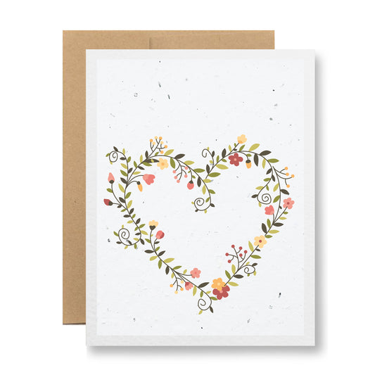 Introducing the "Plantable Greeting Card - {heart wreath of flowers}" by My Store: This charming card showcases a heart shape made from an array of small pink, red, and yellow floral patterns accented with green leaves, all set against a crisp white background. Crafted from eco-friendly handmade seed paper, it comes complete with a partially visible brown envelope.