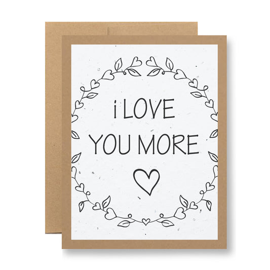 The "I love you more" Plantable Greeting Card by My Store is made from seed paper and features a brown border. The center showcases the phrase "I LOVE YOU MORE," encircled by hand-drawn leaves and hearts, with a small heart below. Once planted, the card releases wildflower seeds that bloom beautifully, combining heartfelt sentiment with environmental sustainability.