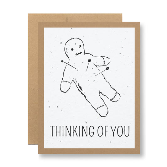 The "Plantable Greeting Card - Thinking of you {voodoo}" by My Store is made from wildflower seed embedded paper and showcases an illustration of a voodoo doll with pins. The phrase "THINKING OF YOU" appears below the image, complemented by a brown envelope.