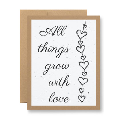 From My Store, the Plantable Greeting Card - "All things grow with love" features elegant script printed on handmade seed paper. Heart shapes are linked by a delicate thread to the right. The card is framed in brown and comes with a matching envelope, offering the delightful promise of blooming wildflower seeds.