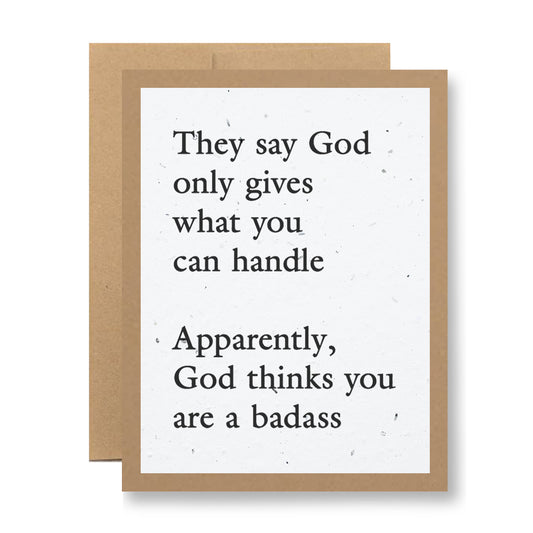 A Plantable Greeting Card from My Store, featuring a brown border and made from wildflower seed-embedded paper, is inscribed with the message: "They say God only gives what you can handle. Apparently, God thinks you are a badass." Behind it is another card that highlights the appeal of biodegradable greeting cards.