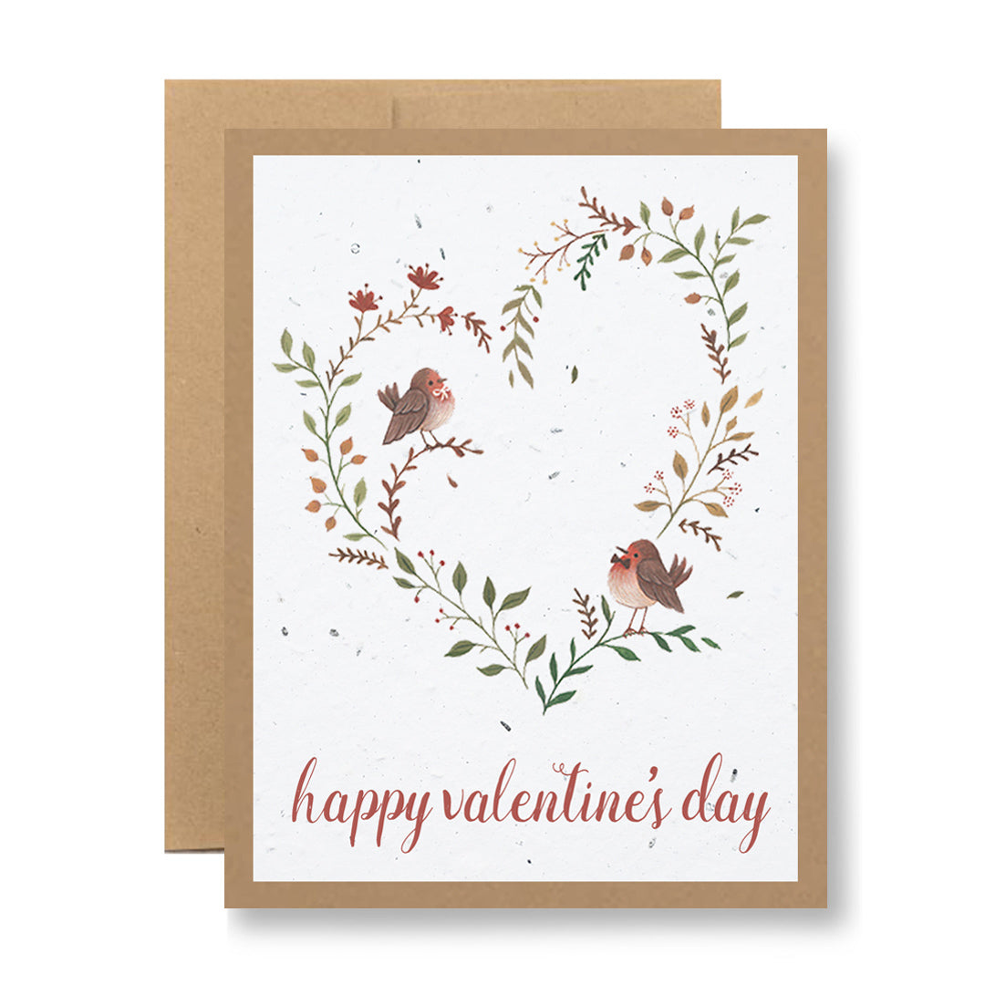 The Plantable Greeting Card - Happy Valentine's Day {birds in a heart} from My Store showcases two birds sitting on a heart-shaped wreath made of green and red leaves. It is created using handmade seed paper, and the words "Happy Valentine's Day" are beautifully written in red cursive at the bottom, surrounded by a brown border.